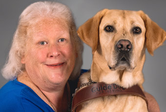 Portrait of Laura Ann wearing a blue shirt next to her Yellow Labrador who is wearing a leather harness with the words Guide Dog for the Blind embossed on the front.
