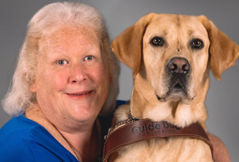 L_Grymes Portrait of Laura Ann wearing a blue shirt next to her Yellow Labrador who is wearing a leather harness with the words Guide Dog for the Blind embossed on the front.